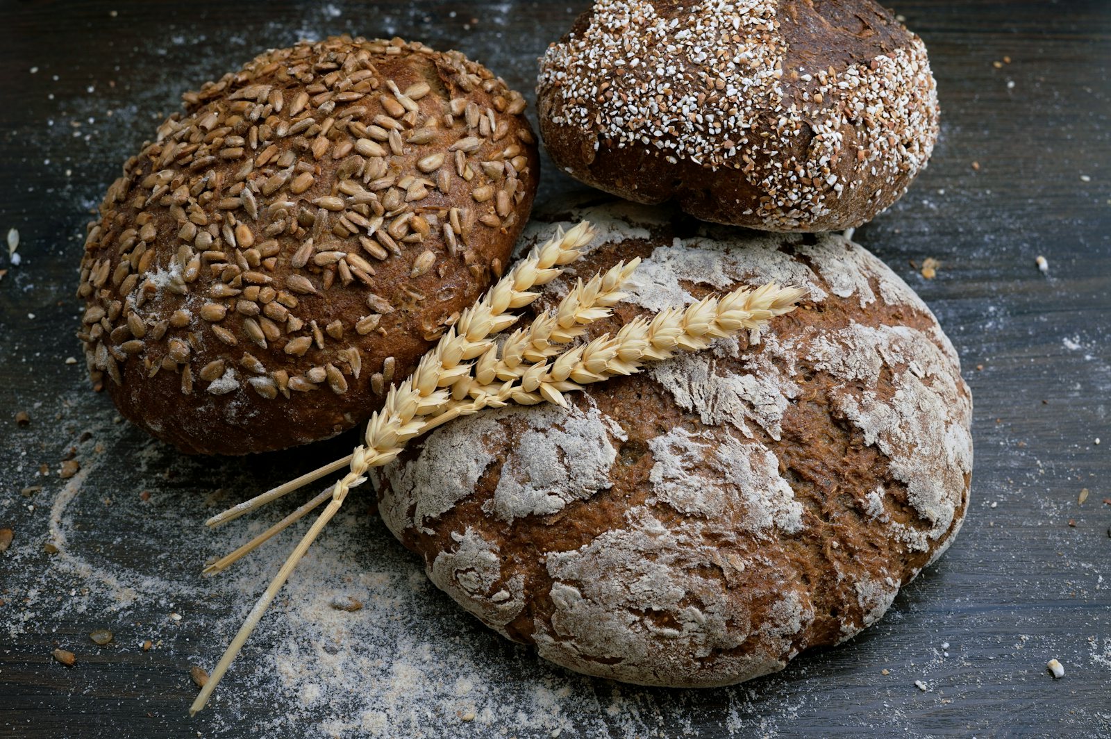 Three seeded bread loaves with wheat stalks on a dark wooden surface.