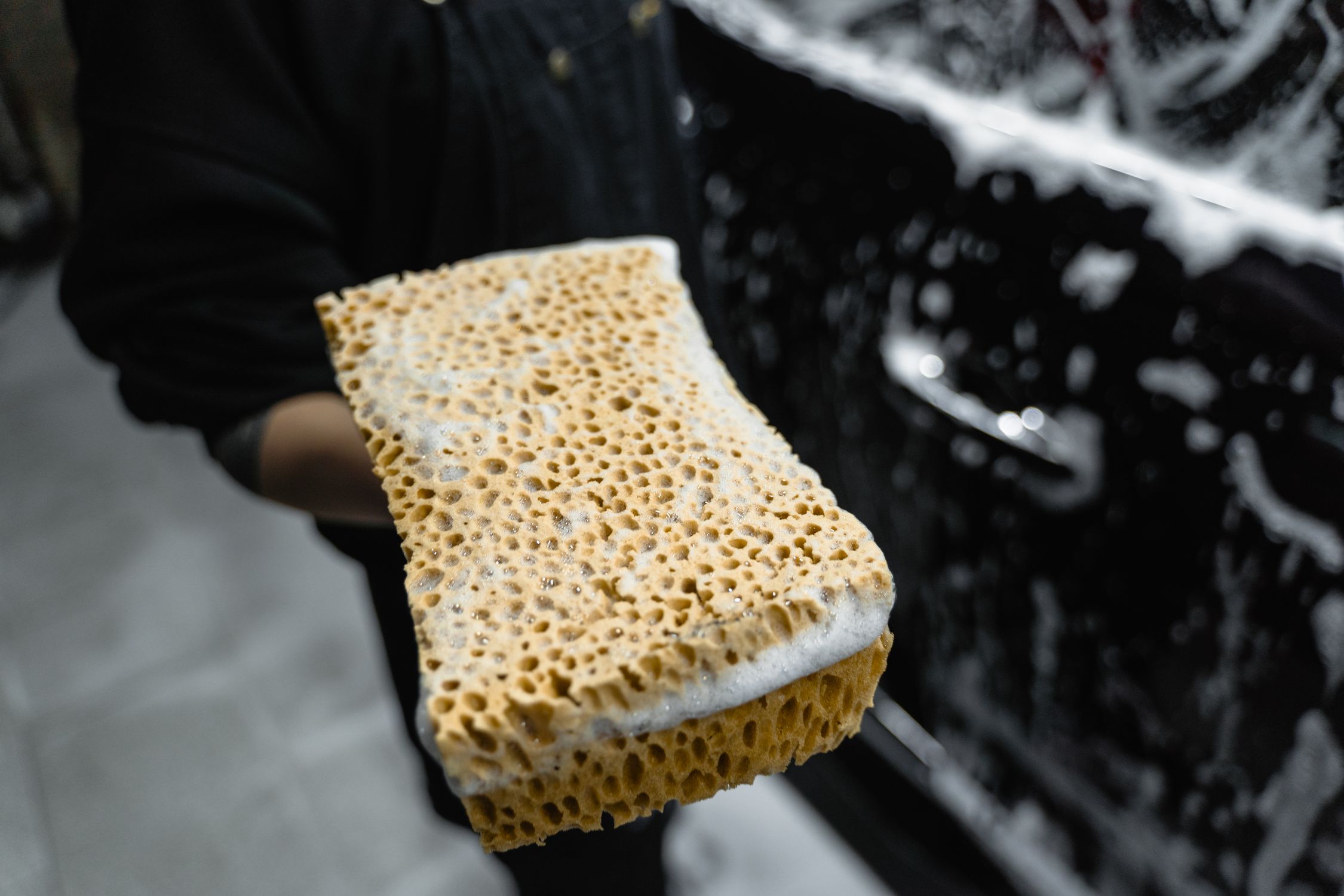 A yellow sponge covered with soap suds held beside a black car.
